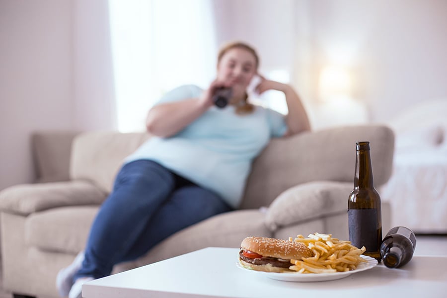 Kobieta z nadwagą siedzi na kanapie, a przed nią stoi piwo i hamburger